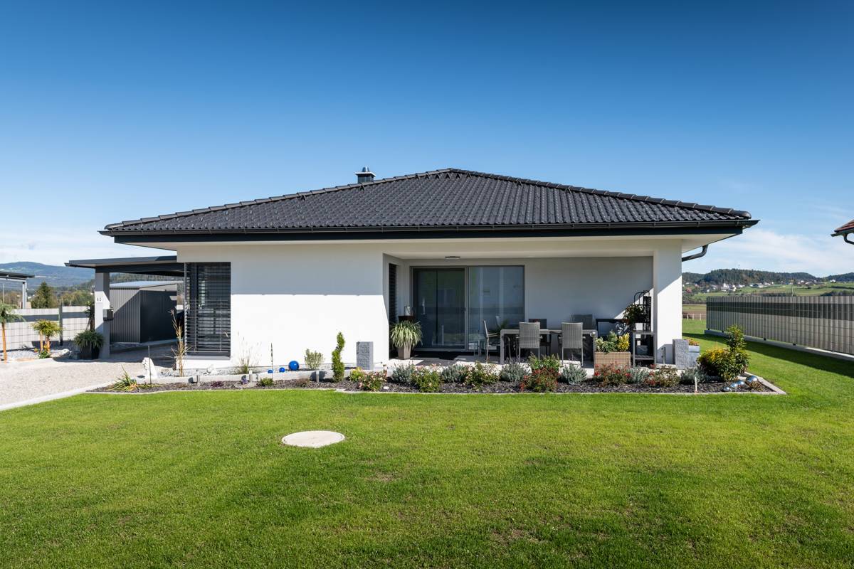 Eingeschoßiger weißer Bungalow mit dunklem Walmdach, überdachter Terrasse und Carport, Gartenansicht