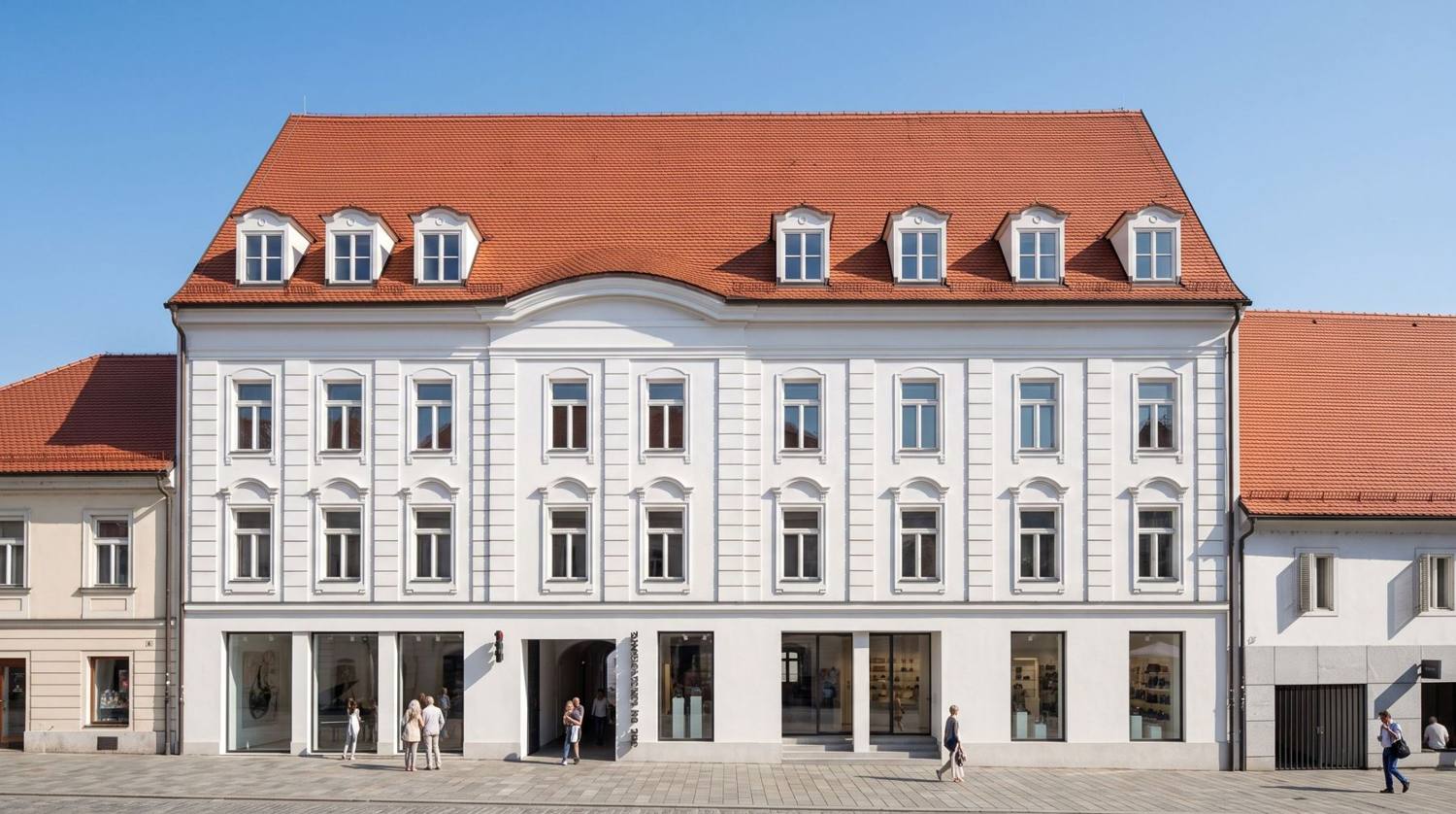 Saniertes barockes Stadthaus mit weißer Stuckfassade, rotem Ziegeldach mit Gauben und Geschäften im Erdgeschoß